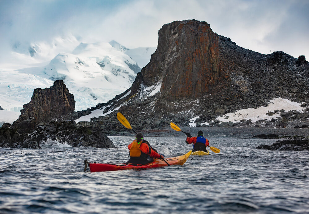 Two kayakers paddle towards rocky outcrop on Antarctic Peninsula